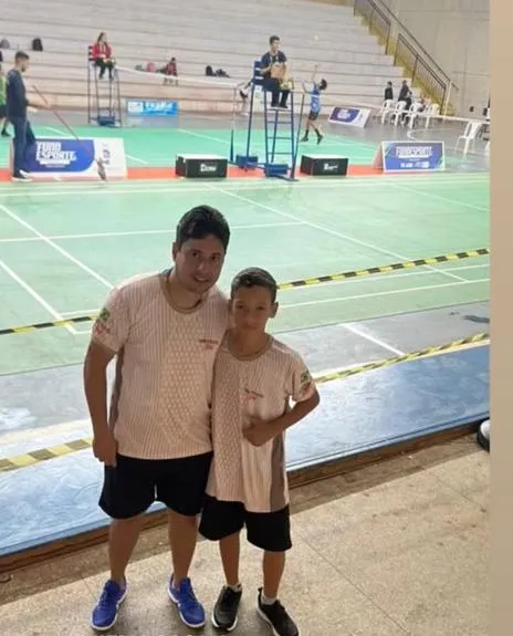 Adulto e menino com camisetas do instituto em um ginásio de badminton.