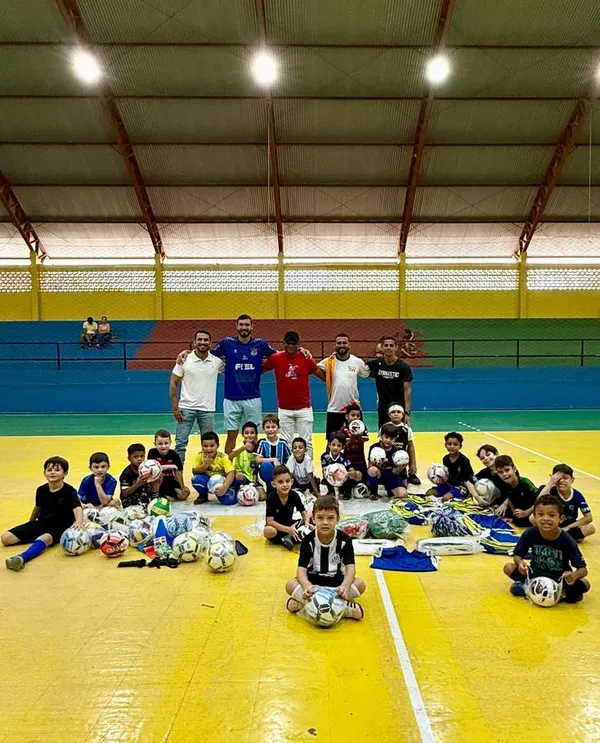 Crianças sentadas na quadra do ginásio com bolas e materiais esportivos recém-entregues.